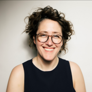 a picture of Oktavia smiling with mid-long curly hair and dark framed glasses. They are wearing a sleeveless dark blue top